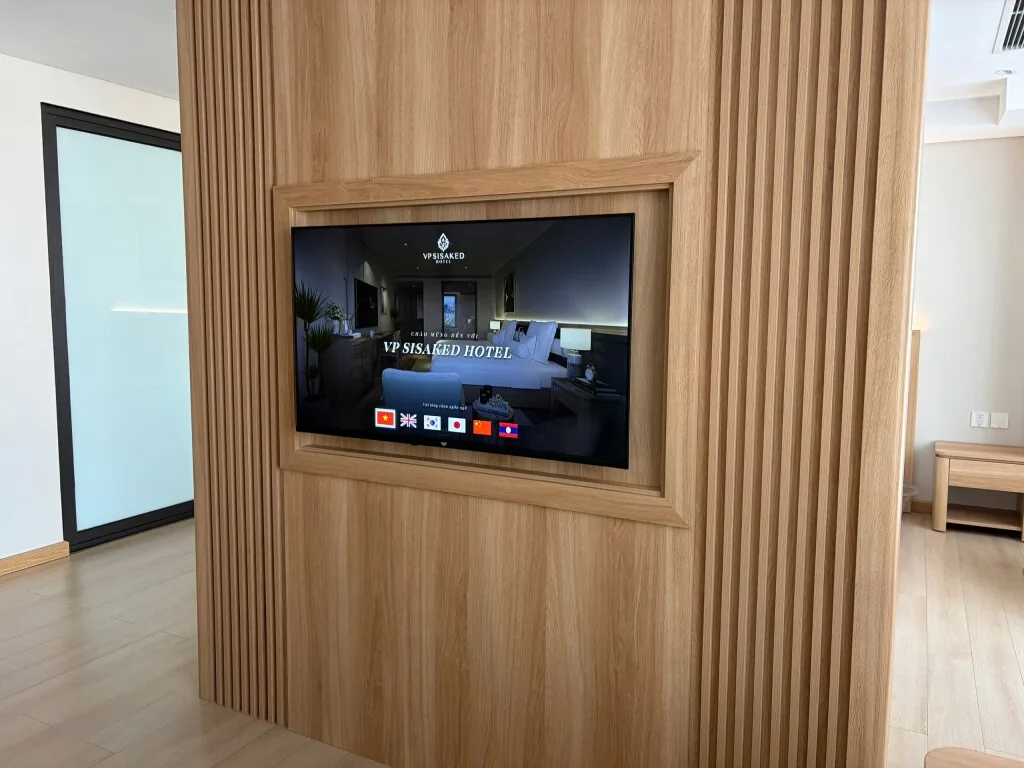 Smart TV gắn trong vách gỗ giữa phòng ngủ VP Sisaked Hotel, không dùng box, màn hình chào đa ngôn ngữ